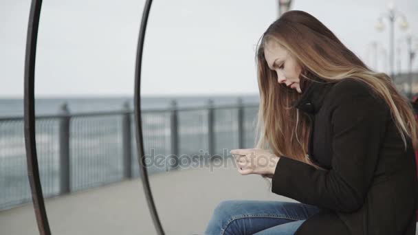 Woman with white smartphone on the bench on the boardwalk overlooking the sea. — Stock Video