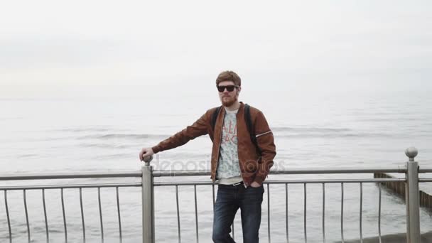 Un joven de moda con barba roja, en una chaqueta de cuero marrón, con gafas de sol Con una mochila, sonríe y mira al mar — Vídeo de stock