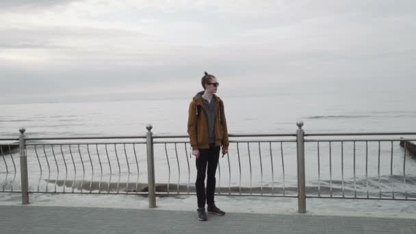 Een modieuze jongeman met, in een bruine jas, dragen van een zonnebril, glimlacht en kijkt naar de zee — Stockvideo