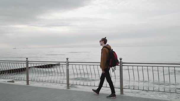 Ein modischer junger Mann mit brauner Jacke, Sonnenbrille, Lächeln und Blick auf das Meer — Stockvideo