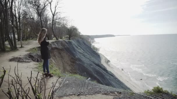 Una joven mira el mar desde una roca, esperando algo. En un abrigo y con el pelo largo, un cielo azul . — Vídeos de Stock