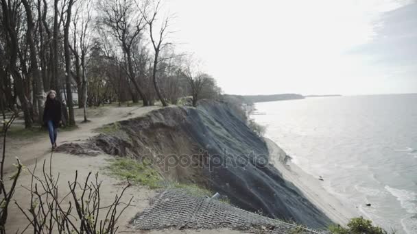 Mladá žena se dívá na moři ze skály, čekání na něco. V kabátě a s dlouhými vlasy, modrá obloha. — Stock video