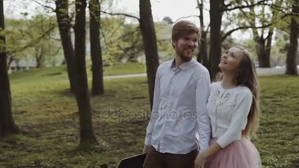 Молода пара закохана у прогулянку в літньому парку, тримаючись за руки. Жінка в рожевій спідниці tutu чоловік з дошкою в руках тривісний стабілізований відстеження дріб — стокове відео