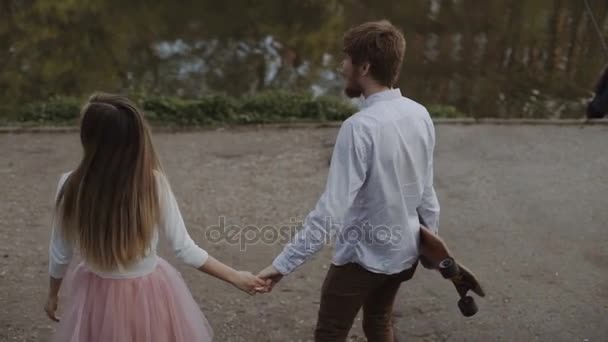 Young couple in love walking in summer park holding hands. Woman in pink tutu skirt man with longboard in hands Three-axis Gimbal stabilized tracking shot — Stock Video