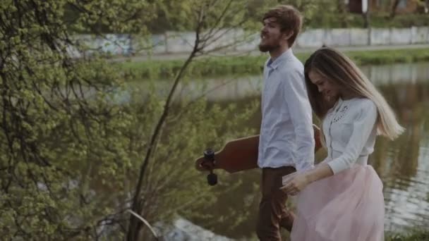 Pareja joven enamorada caminando en el parque de verano tomados de la mano. Mujer en falda tutú rosa hombre con longboard en las manos Gimbal de tres ejes estabilizado tiro de seguimiento — Vídeo de stock