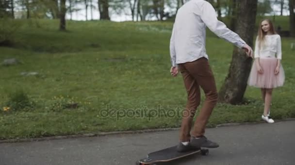 Pareja joven enamorada caminando en el parque de verano tomados de la mano. Mujer en falda tutú rosa hombre con longboard en las manos Gimbal de tres ejes estabilizado tiro de seguimiento — Vídeo de stock