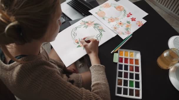 Eine junge Künstlerin malt mit Aquarell einen Wasserpinsel, Blumen, einen Kranz aus grünen Blättern. Nahaufnahme — Stockvideo