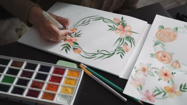 Una joven artista, pinta con acuarela pinta flores de pincel de agua, una corona de hojas verdes. de cerca — Vídeo de stock