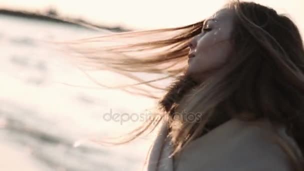 Mujer joven con el pelo largo vestida con una túnica transparente posando al atardecer en la playa — Vídeos de Stock