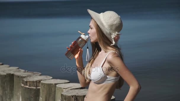 Güneş banyo seksi bir genç kadın bir yaz şapka ve beyaz bikini mayo suyu şişeden içer.. — Stok video