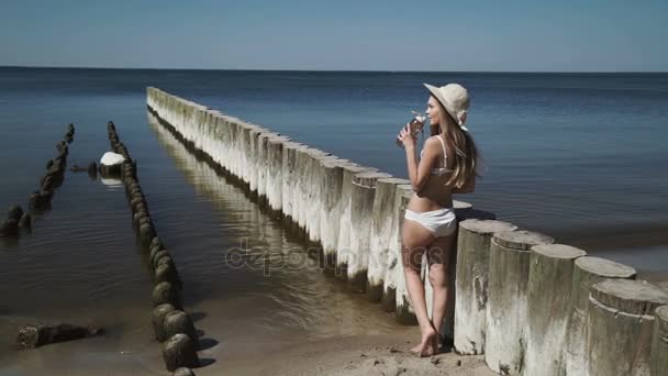 Una joven sexy en un sombrero de verano y un traje de baño de bikini blanco, tomando baños de sol, bebe agua de una botella . — Vídeos de Stock