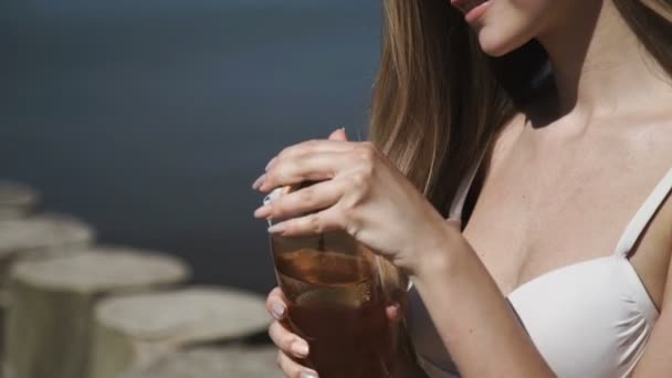 Una joven sexy en un sombrero de verano y un traje de baño de bikini blanco, tomando baños de sol, bebe agua de una botella . — Vídeos de Stock
