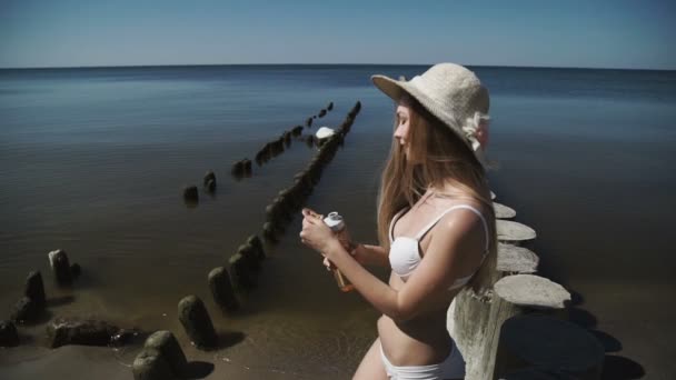 En sexig ung kvinna i en sommar hatt och en vit bikini baddräkt, med sol bad, dricker vatten ur en flaska. — Stockvideo