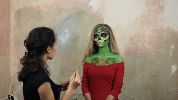 Woman artist doing aquagrim face art on halloween tassels green scary glamorous rhinestones skeleton. Mexican Princess Sugar Skull. woman model posing in front of camera in red dress. — Stock Video