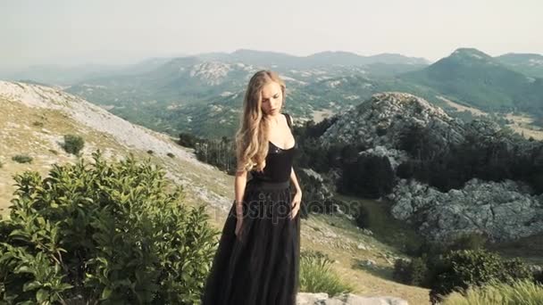 Bella giovane donna modello in un nero soffice elegante abito lungo in posa sulla macchina fotografica sullo sfondo di un paesaggio di montagna . — Video Stock