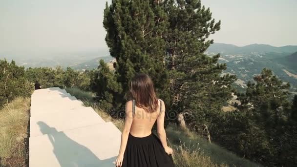 Hermosa modelo de mujer joven con el pelo largo en un vestido largo elegante esponjoso negro está caminando a lo largo del balcón en el fondo de un paisaje de montaña. vista de verano de las montañas . — Vídeos de Stock