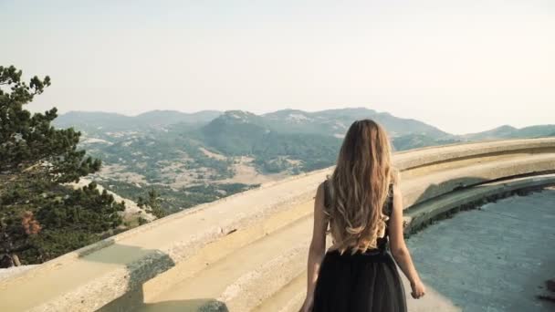 Hermosa modelo de mujer joven con el pelo largo en un vestido largo elegante esponjoso negro está caminando a lo largo del balcón en el fondo de un paisaje de montaña. vista de verano de las montañas . — Vídeos de Stock