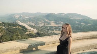 güzel genç kadın modeli siyah kabarık zarif uzun elbise uzun saçlı bir dağ manzarası arka balkonda boyunca yürüyor. dağlarda yaz görünümü.