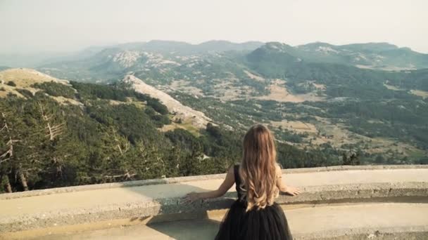 Hermosa modelo de mujer joven con el pelo largo en un vestido largo elegante esponjoso negro está caminando a lo largo del balcón en el fondo de un paisaje de montaña. vista de verano de las montañas . — Vídeos de Stock