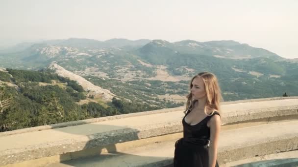 Το μοντέλο όμορφη νεαρή γυναίκα με μακριά μαλλιά με ένα μαύρο χνουδωτό κομψό μακρύ φόρεμα είναι το περπάτημα κατά μήκος το μπαλκόνι στο φόντο ένα ορεινό τοπίο. καλοκαίρι θέα προς τα βουνά. — Αρχείο Βίντεο