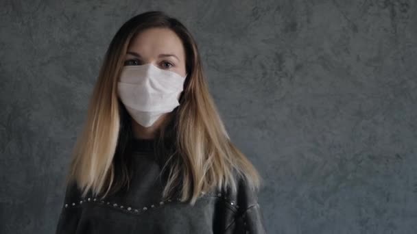 Giovane donna indossa una maschera medica protettiva sul viso contro il muro grigio — Video Stock