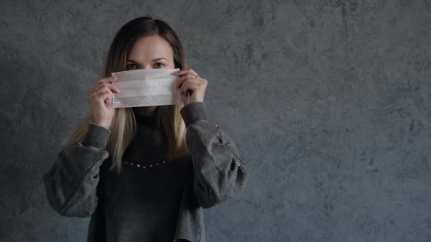 Giovane donna indossa una maschera medica protettiva sul viso contro il muro grigio — Video Stock