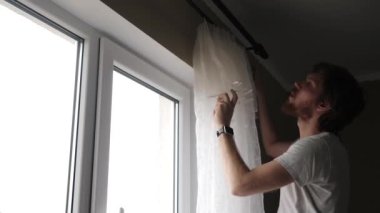 Genç bir adam pencereye perde asıyor.