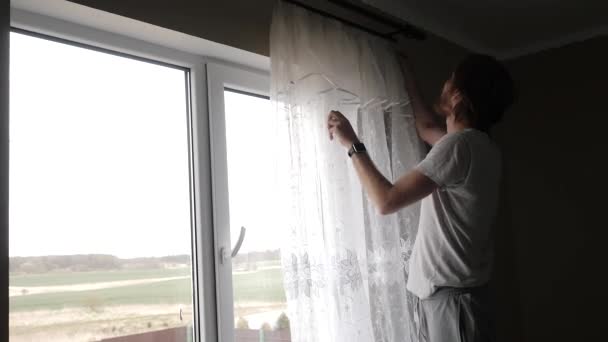 Um jovem pendura cortinas na janela — Vídeo de Stock