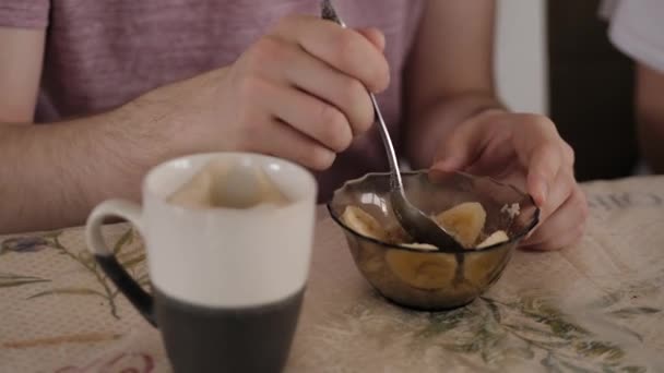 Jovem de óculos come mingau de cereal com banana para café da manhã e bebe café — Vídeo de Stock