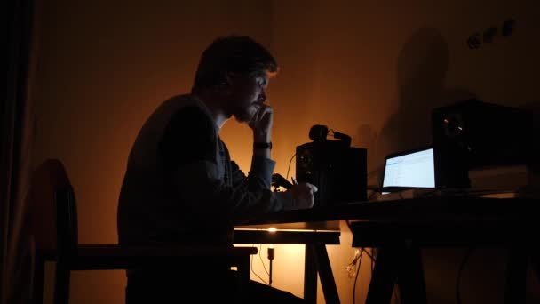 En ung man sitter vid en bärbar dator på natten och skriver i en anteckningsbok — Stockvideo