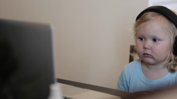 Маленькая девочка в больших наушниках сидит за ноутбуком и смотрит мультики — стоковое видео