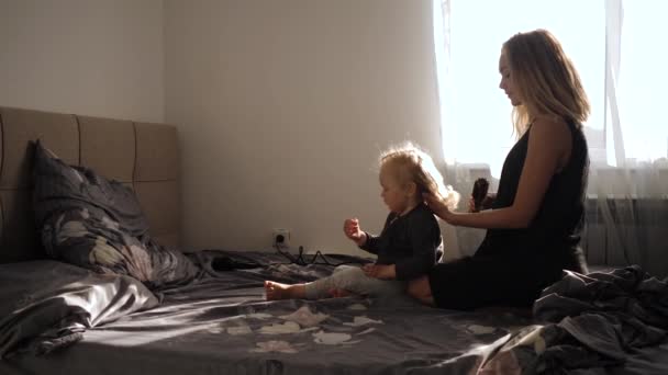 En ung mamma gör frisyr liten flicka hemma sitter på sängen — Stockvideo