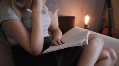 Genç bir kadın kitap okuyor. Evdeki kanepede oturuyor.