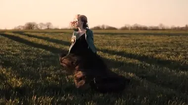 Uzun siyah elbiseli ve deri ceketli genç bir kadın tarlada koşuşturuyor.