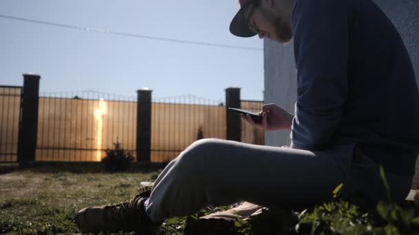 Jovem estudante do sexo masculino sentado na grama e olhando para smartphone exalando fumaça — Vídeo de Stock