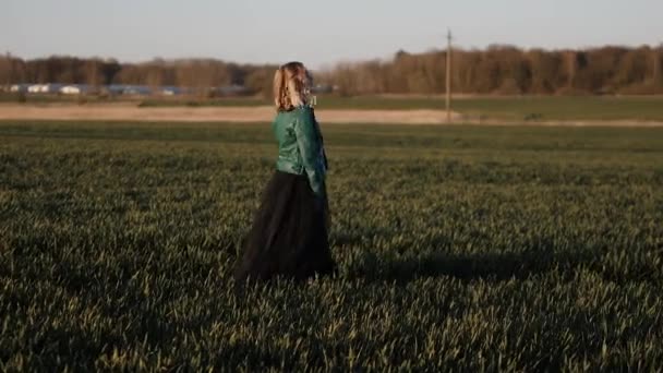 Mladá žena v dlouhých černých šatech a kožené bundě pobíhající po poli — Stock video