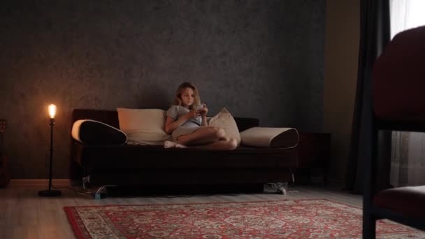 Νεαρή γυναίκα κάθεται στον καναπέ στο σαλόνι με τηλέφωνο στο χέρι — Αρχείο Βίντεο