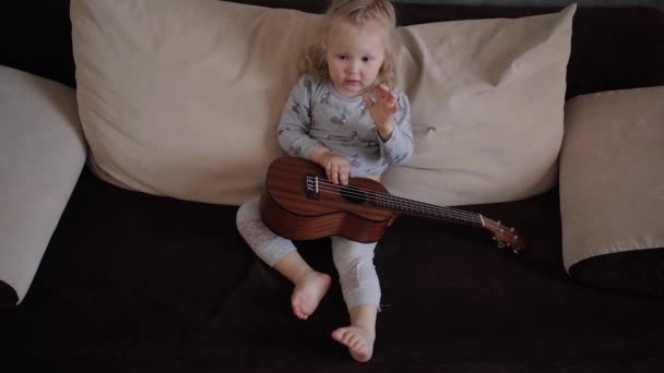 Uma menina aprende a tocar um ukulele instrumento musical no sofá — Vídeo de Stock