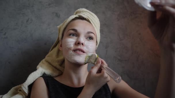 Una mujer joven obtiene crema belleza máscara blanca un cepillo en la cara — Vídeo de stock