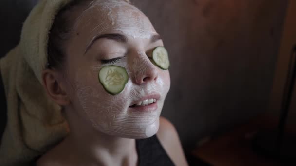 Una giovane donna ottiene una maschera facciale bianca con cetrioli cura della casa divertente — Video Stock
