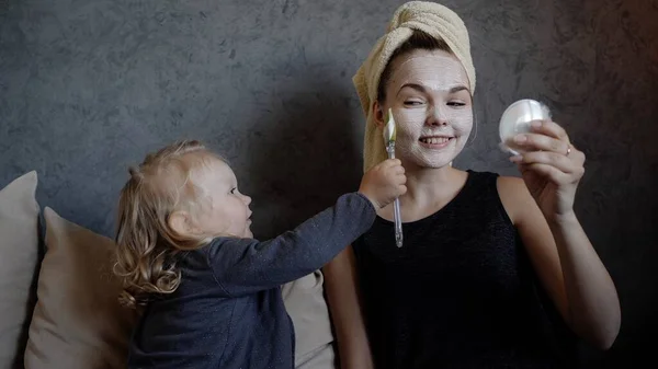 Jong moeder en dochtertje met een wit masker met komkommers op haar gezicht — Stockfoto
