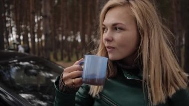 Genç bir kadın ormanda sıcak çay içiyor ve lezzetli bir sandviç yiyor.
