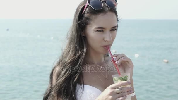 Красивая девушка пьет коктейль мохито на фоне моря — стоковое видео