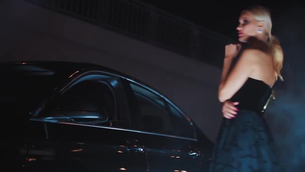 Красива блондинка позує в чорній сукні вночі біля машини — стокове відео