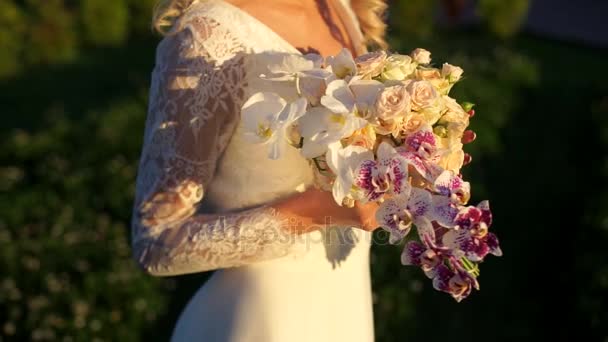 Die Braut berührt mit ihren Händen einen Strauß Rosen und Orchideen in den hellen Strahlen der untergehenden Sonne. — Stockvideo