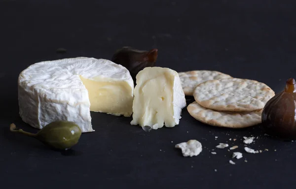 Camembert cheese, figs, caper berry and crackers on black backgr — Stock Photo, Image