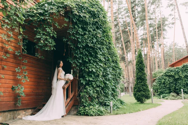 Portrait of the bride in a wedding dress with a bouquet of flowers at the entrance to a wooden house, overgrown with greenery.