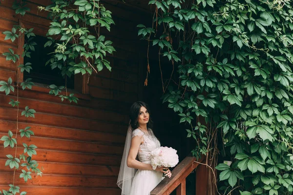 Portrait of the bride in a wedding dress with a bouquet of flowers at the entrance to a wooden house, overgrown with greenery.