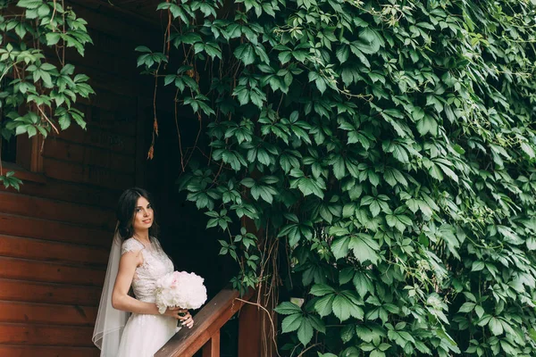 Portrait of the bride in a wedding dress with a bouquet of flowers at the entrance to a wooden house, overgrown with greenery.