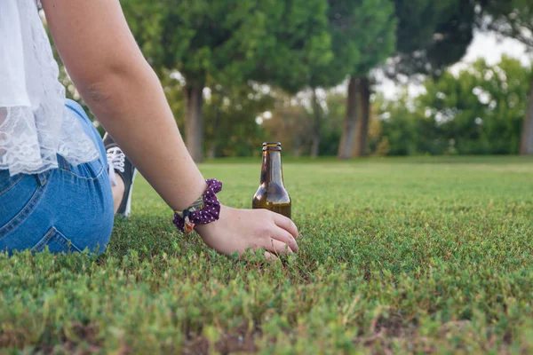 Menina tem sua garrafa de cerveja no chão, enquanto olha para o horizonte pensativo. Sua mão e cerveja descansando na grama . — Fotografia de Stock